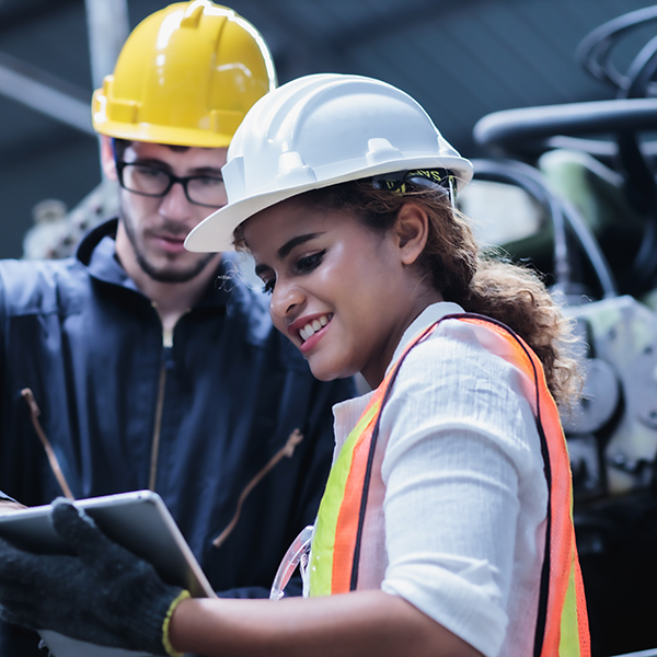 Two apprentices wearing safety helmets review information on a tablet in an industrial environment as part of a work-study training program